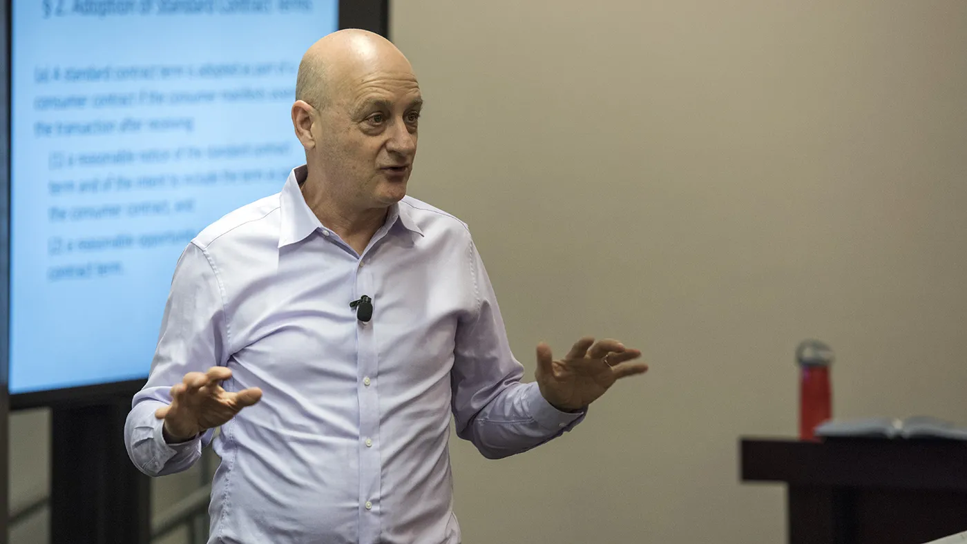Omri Ben-Shahar gestures in front of a classroom with slides on a display screen behind him.