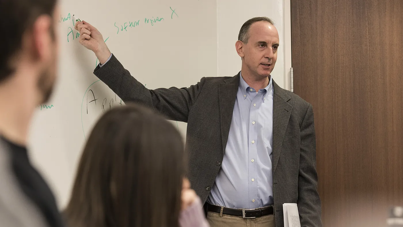 Eric Posner, in a blue shirt and grey sportscoat, points at the white board behind him.
