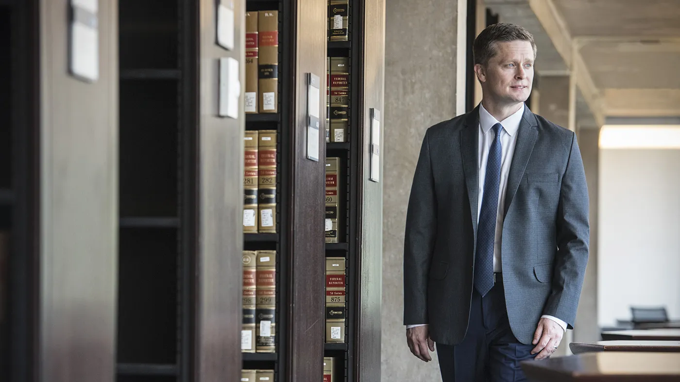Anthony Casey, in a blue suit, white shirt, and blue tie, walks among the library stacks.