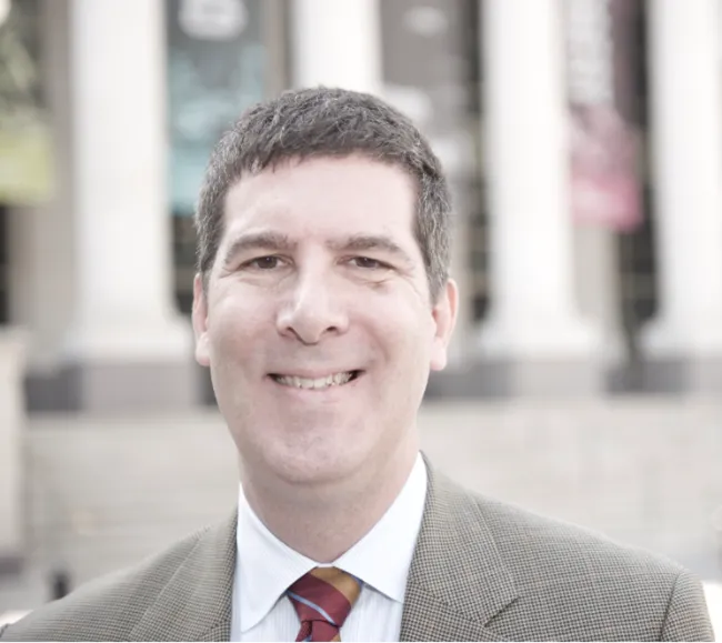 In a suit and red tie, Bob Mendes stands outside a building with columns.