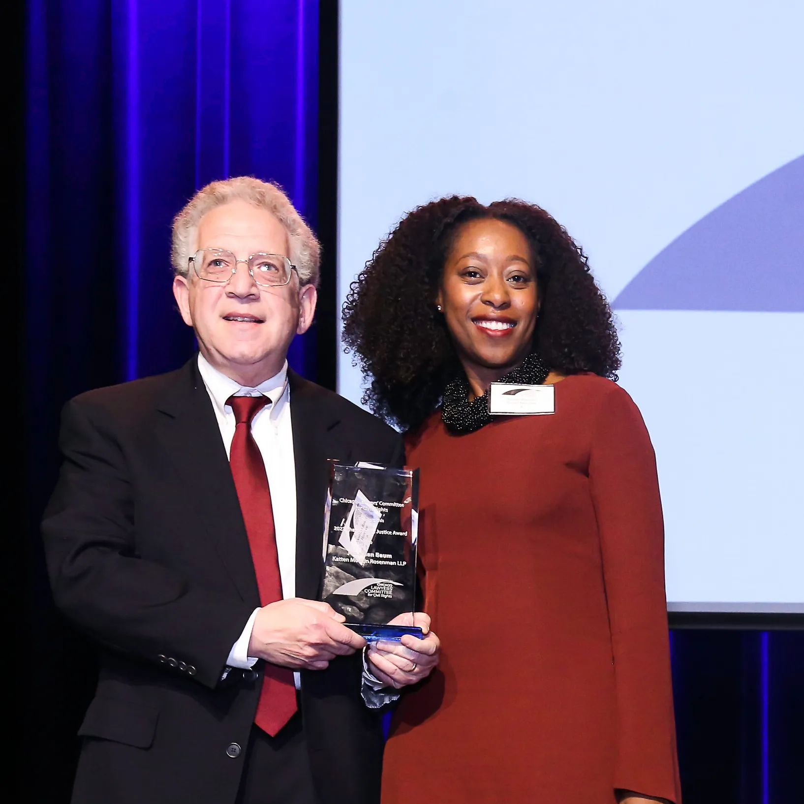 In a suit and tie, Jonathan Baum stands on a stage with a woman in a red dress. The screen behind them reads, "Chicago Lawyer's Committee for Civil Rights"